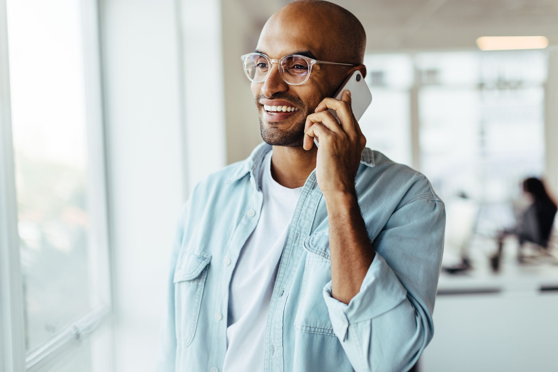 Man with glasses smiles while talking on a phone near a window in an office.