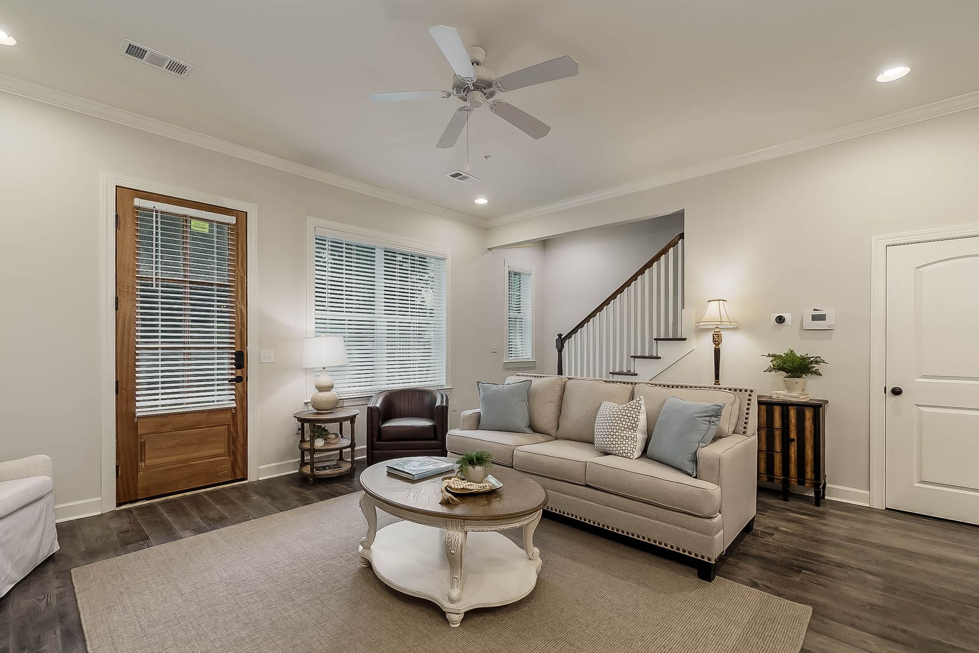 Living room with sofa, coffee table, rug, and dark wood flooring. Stairs lead upward, door to the side.