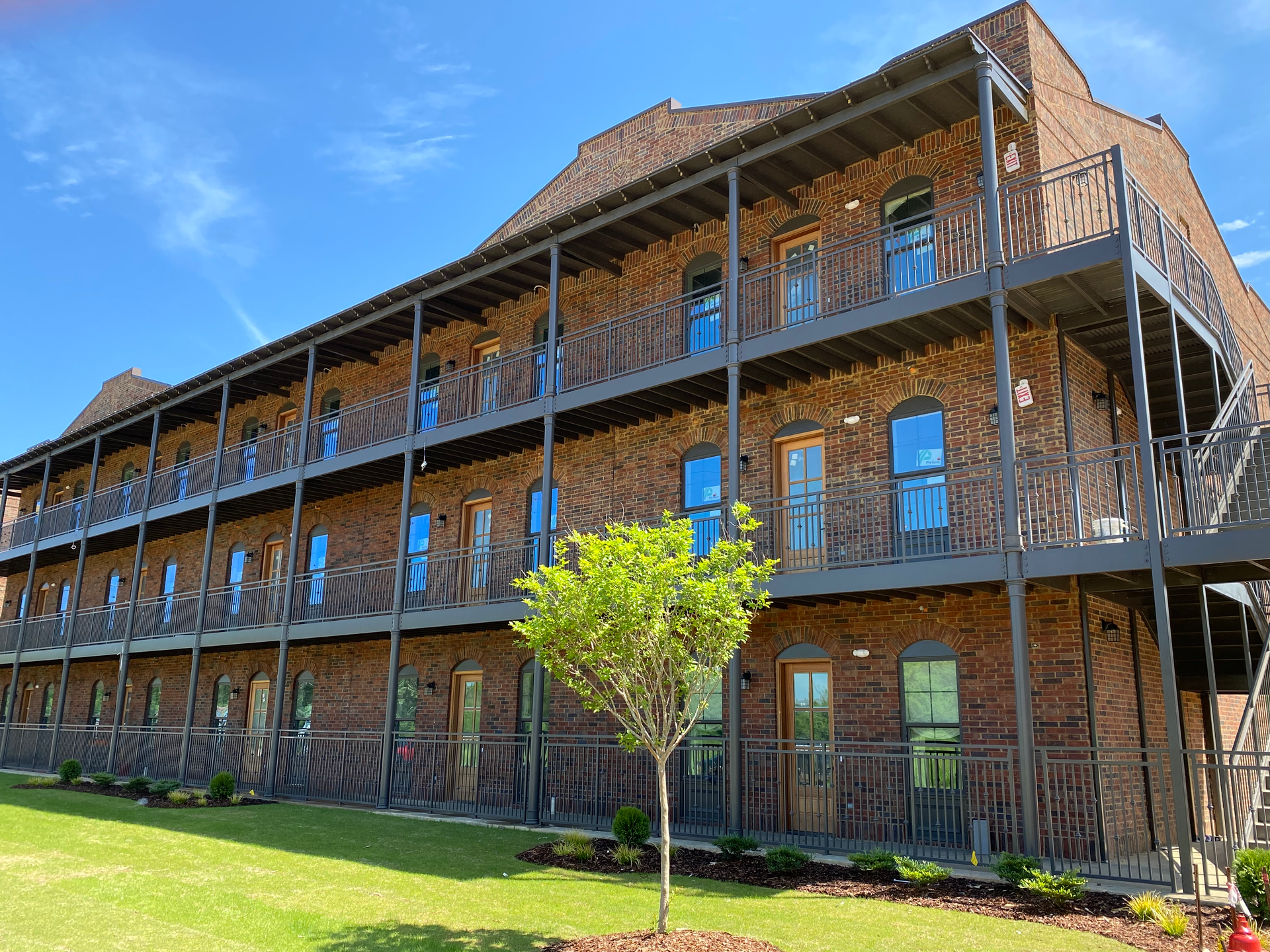 Three-story brick building with multiple balconies, blue sky, and a small tree in front.