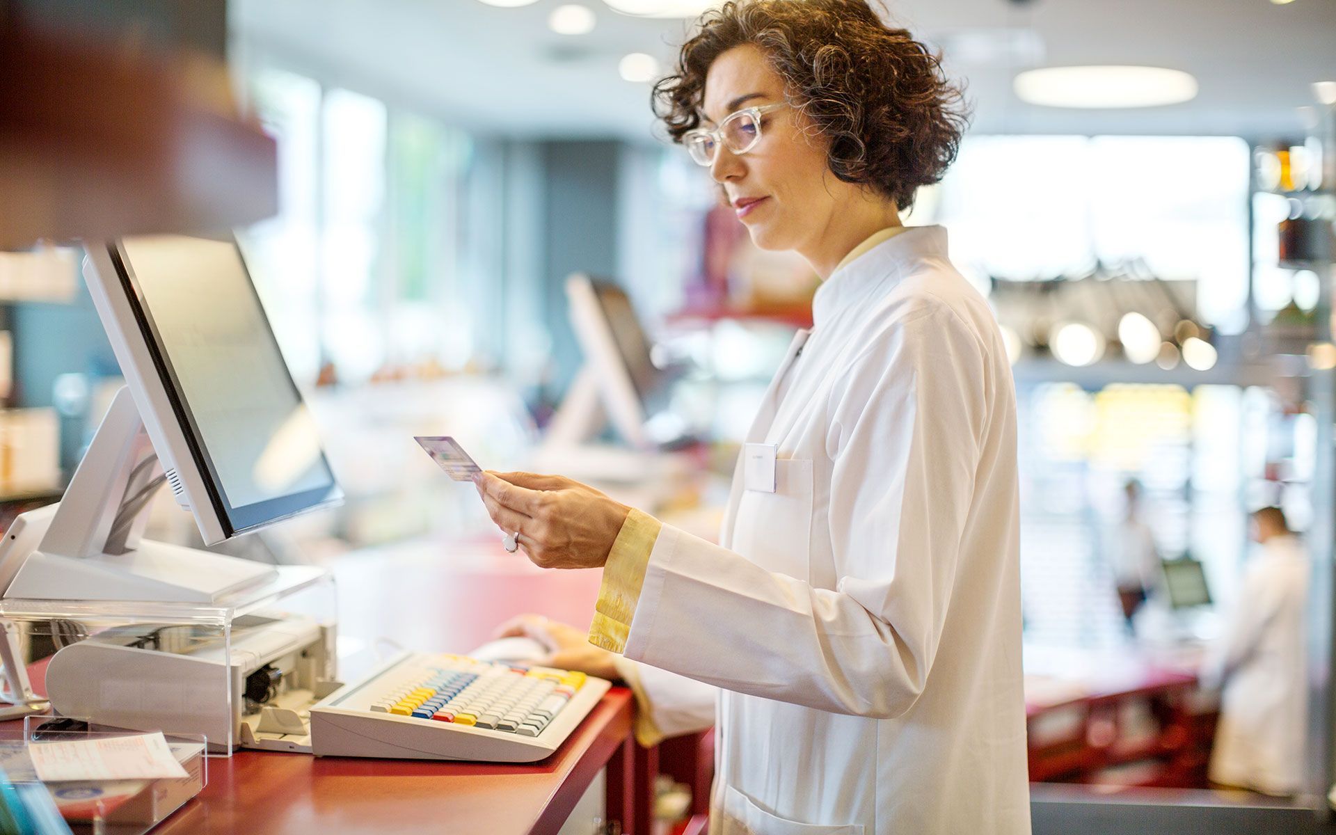 Pharmacist in a white coat, handling a credit card at a pharmacy counter.