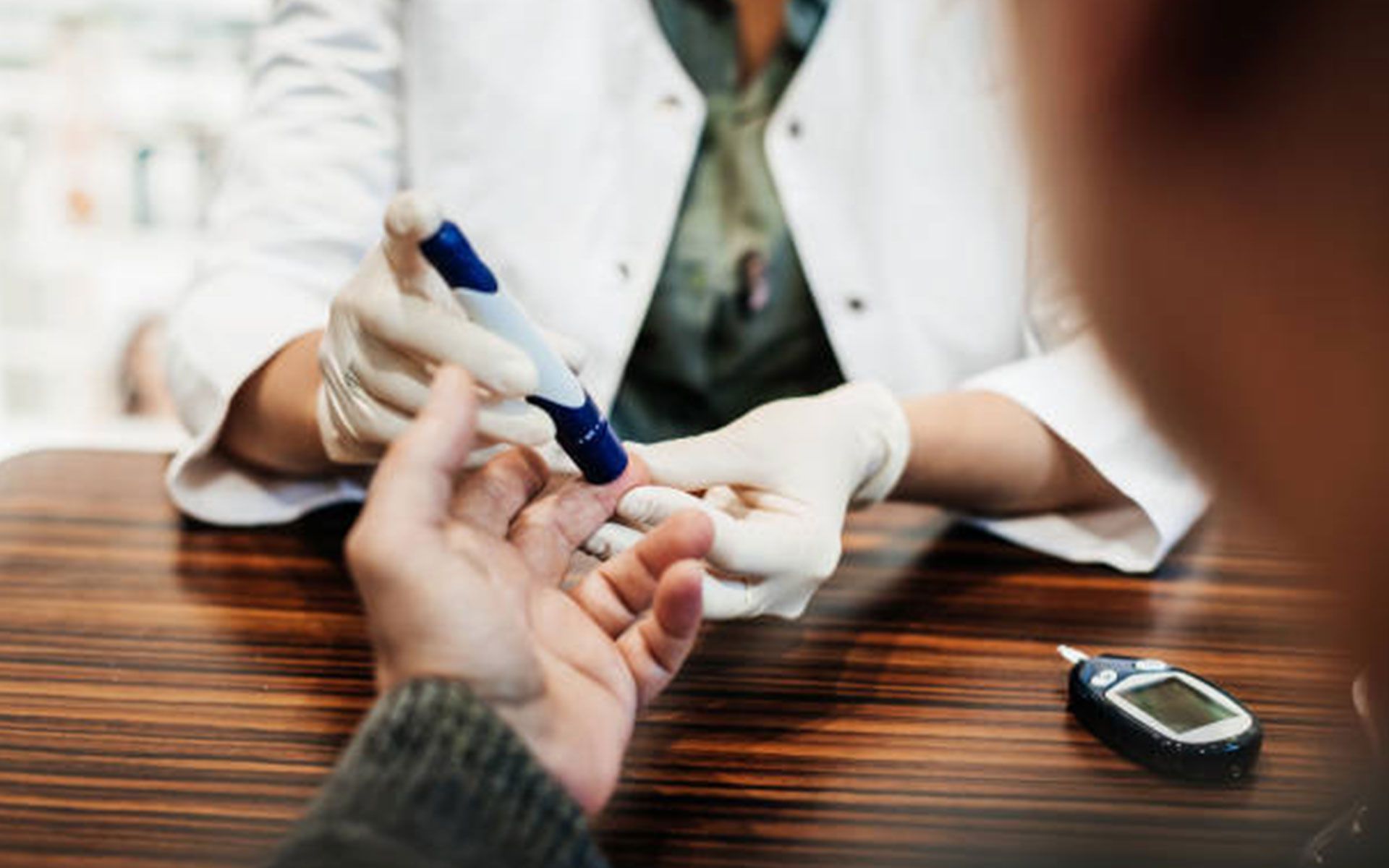 A healthcare worker wearing gloves pricks a patient's finger with a lancet to test blood sugar levels.
