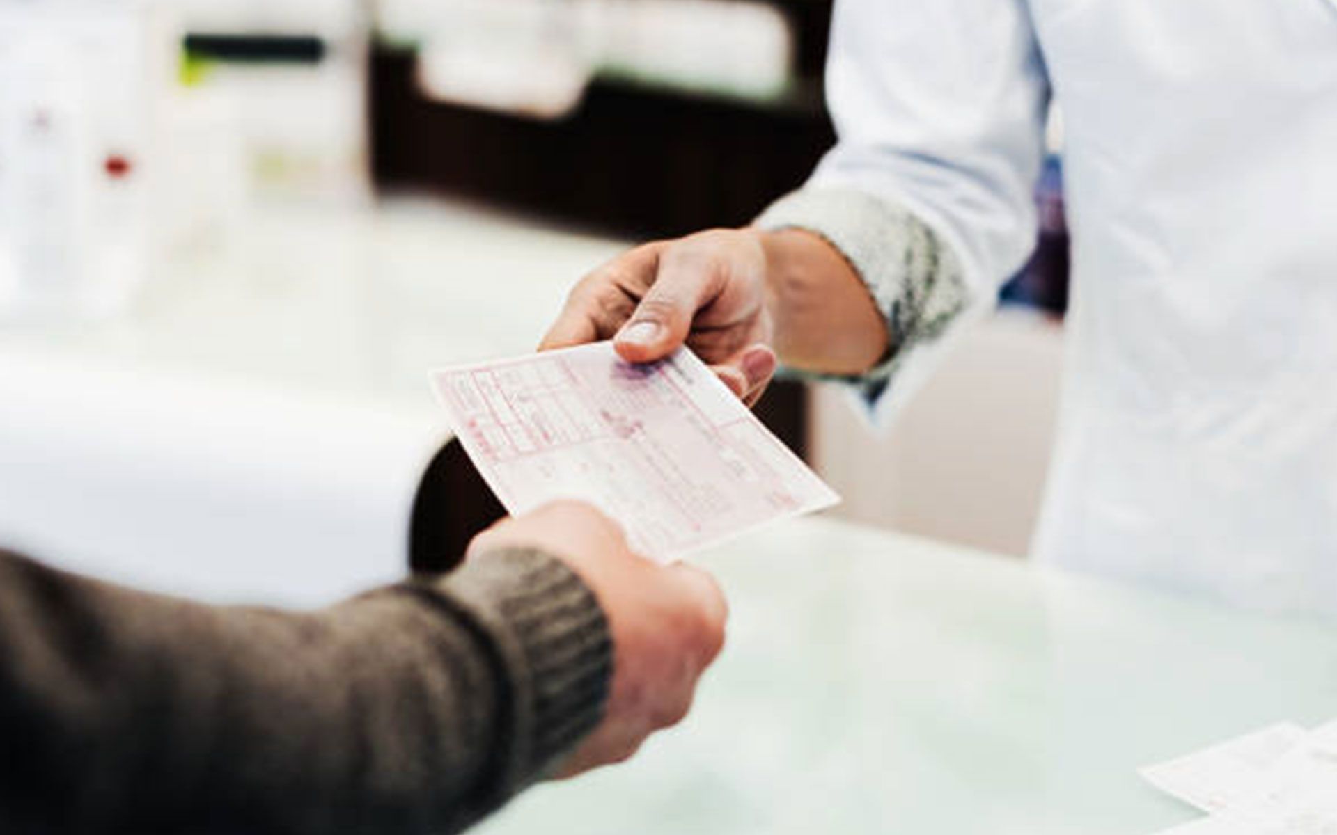 Person in white coat handing prescription to another person over a counter.
