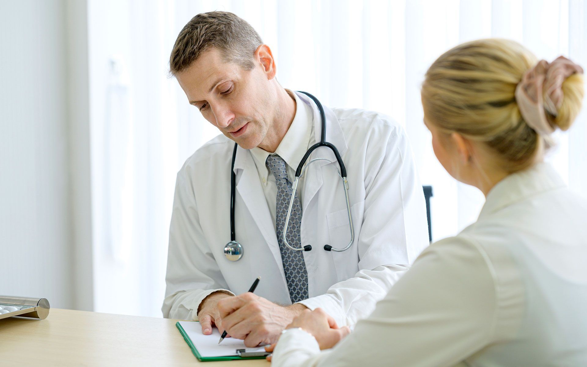 Doctor in white coat writing notes, talking with a patient at a desk.