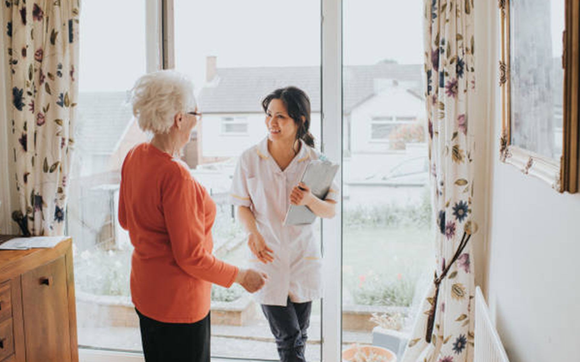 Elderly woman greets a care worker at a doorway, both smiling. White uniform, home interior.