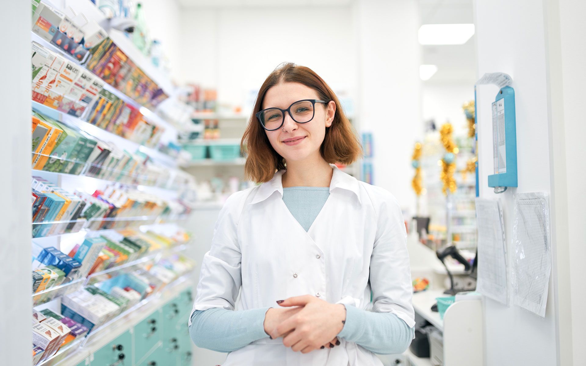 Pharmacist in white coat smiles in a well-stocked pharmacy, wearing glasses.