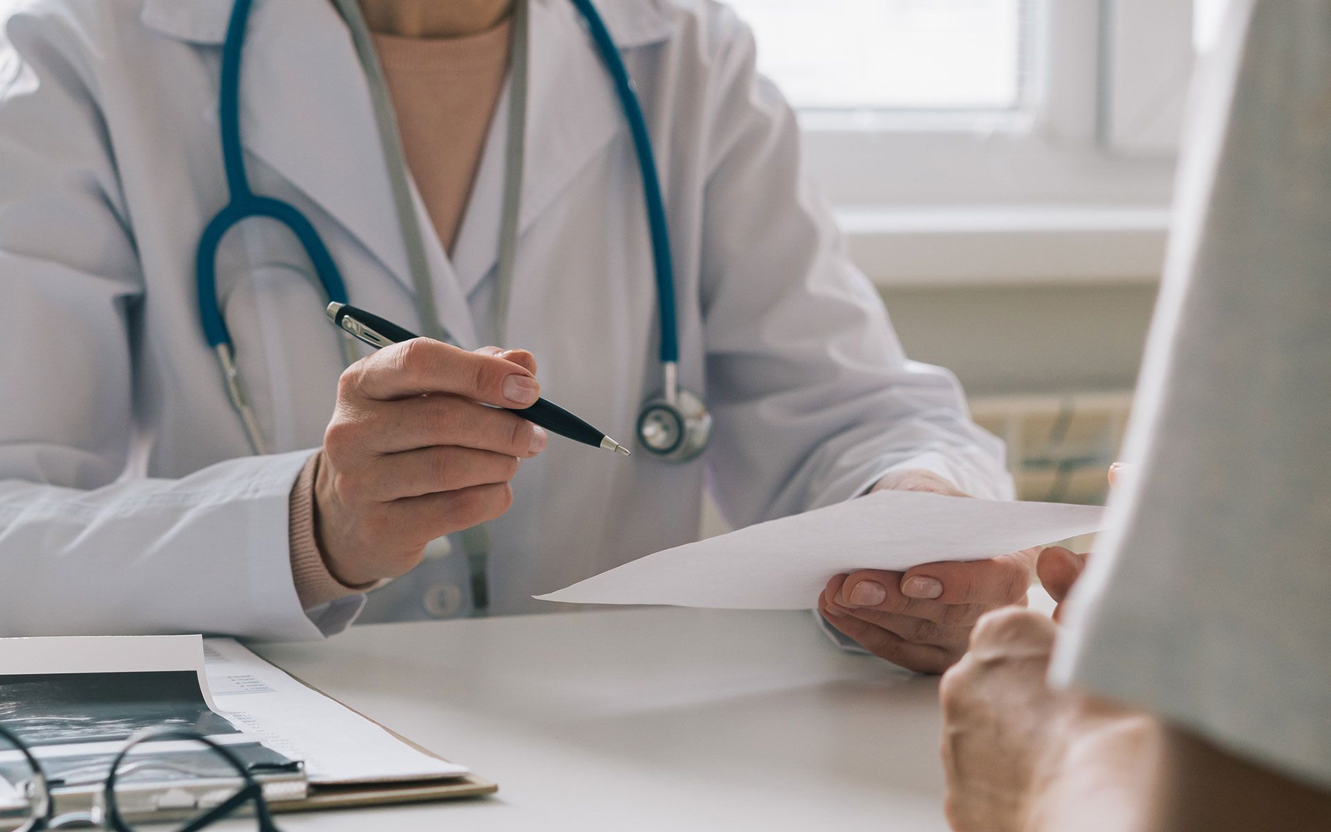 Doctor handing a patient a prescription at a desk. A stethoscope hangs around the doctor's neck.