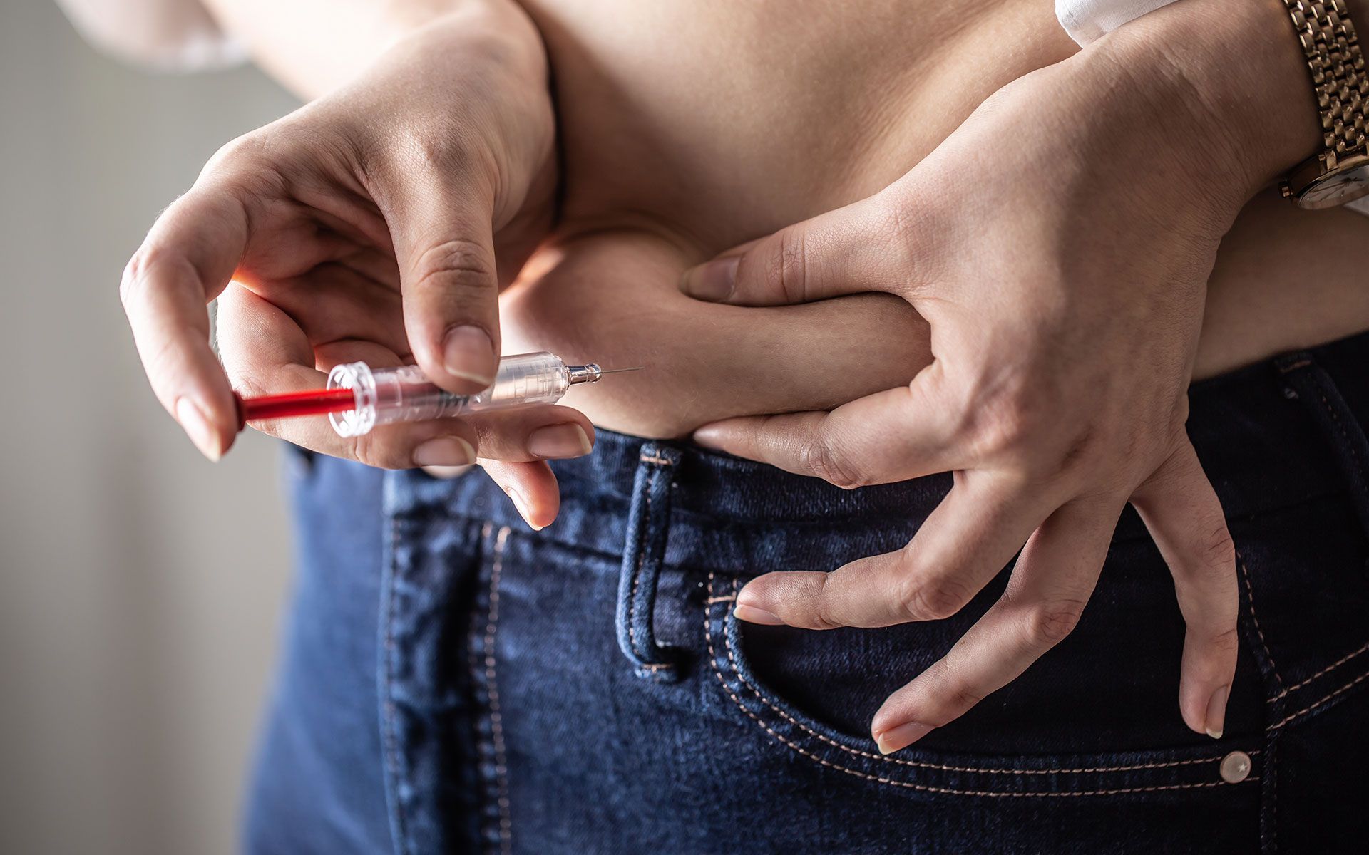 Person injecting insulin into abdominal fat, wearing blue jeans.
