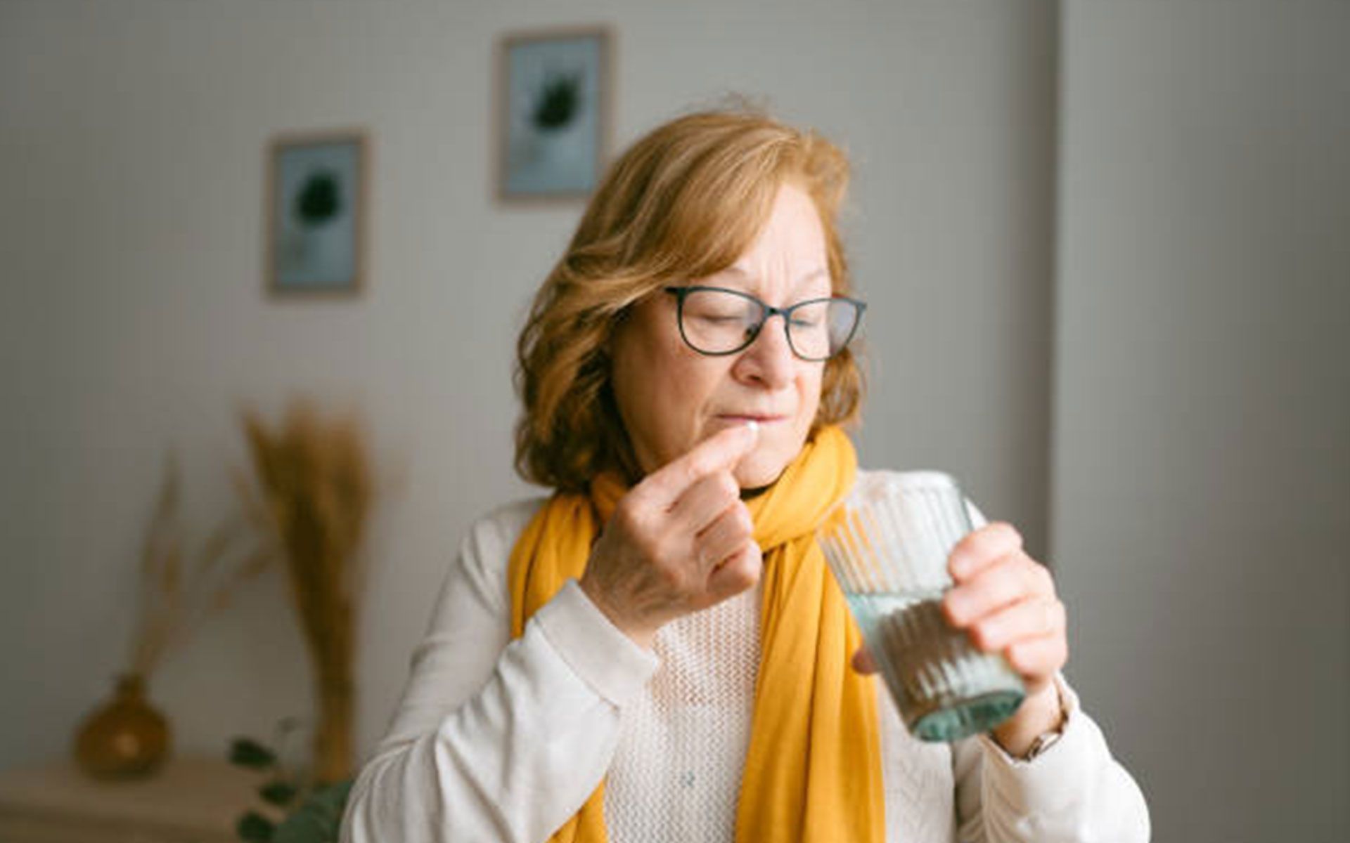 Older woman with glasses takes a pill with water. She is wearing a yellow scarf indoors.