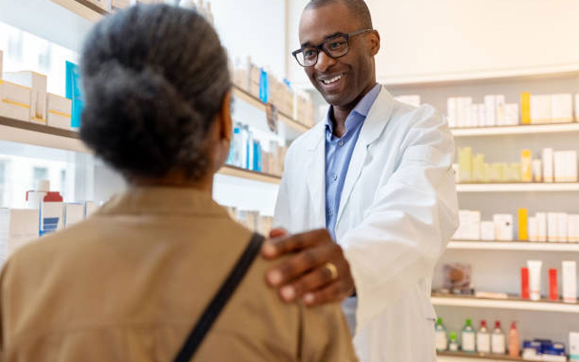 Pharmacist with hand on customer's shoulder, smiling in pharmacy, shelves of products in background.