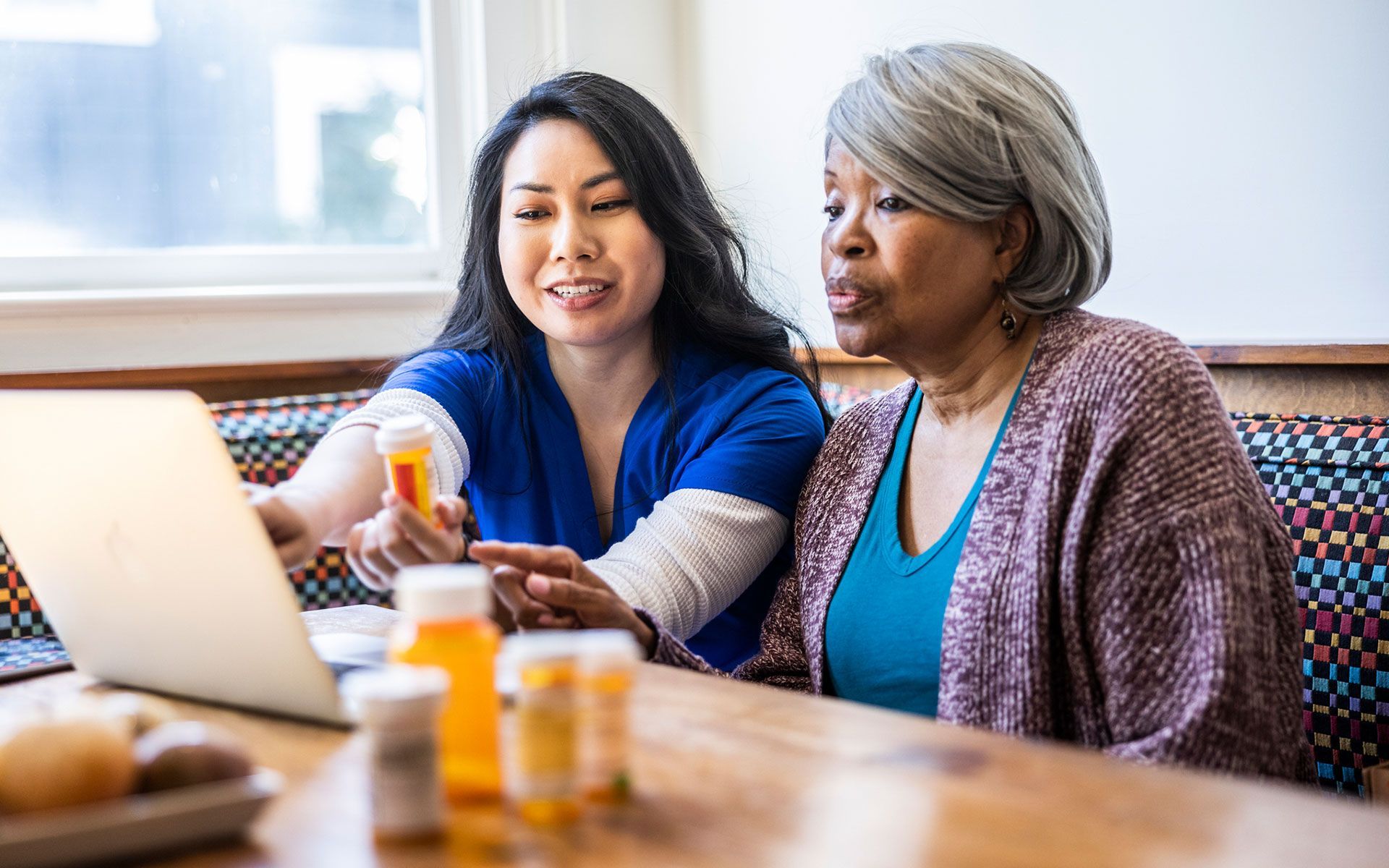 A healthcare provider shows a senior woman medication info on a laptop. Both are looking at the screen at a table.