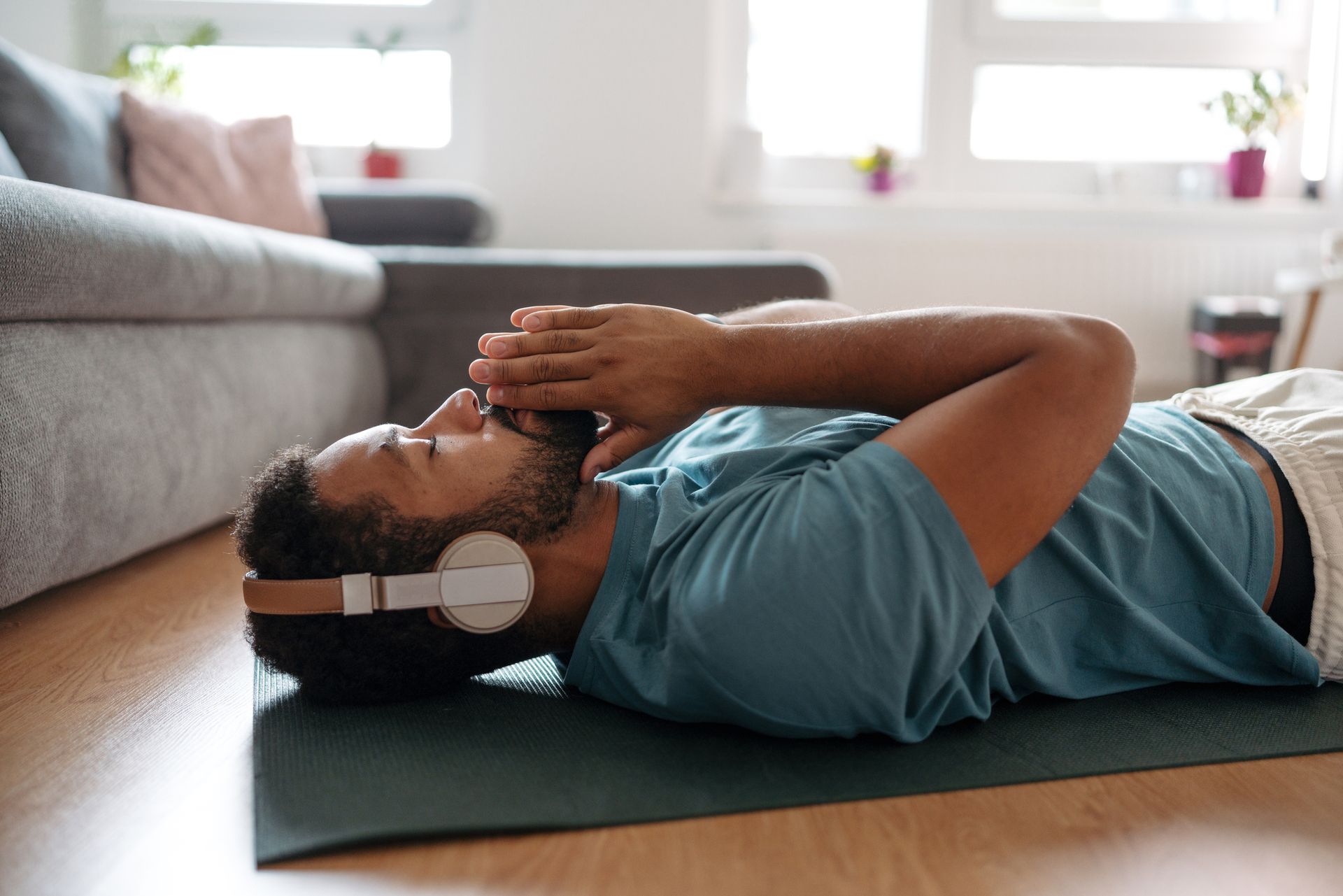 Man lying on a yoga mat, eyes closed, hands folded, wearing headphones, in a living room.