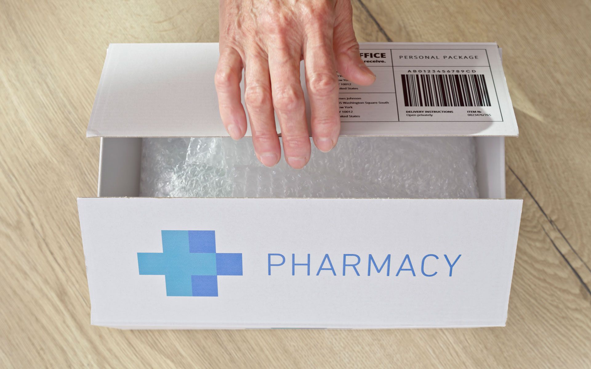 Hand opening a pharmacy delivery box on wooden floor. The box contains wrapped items.