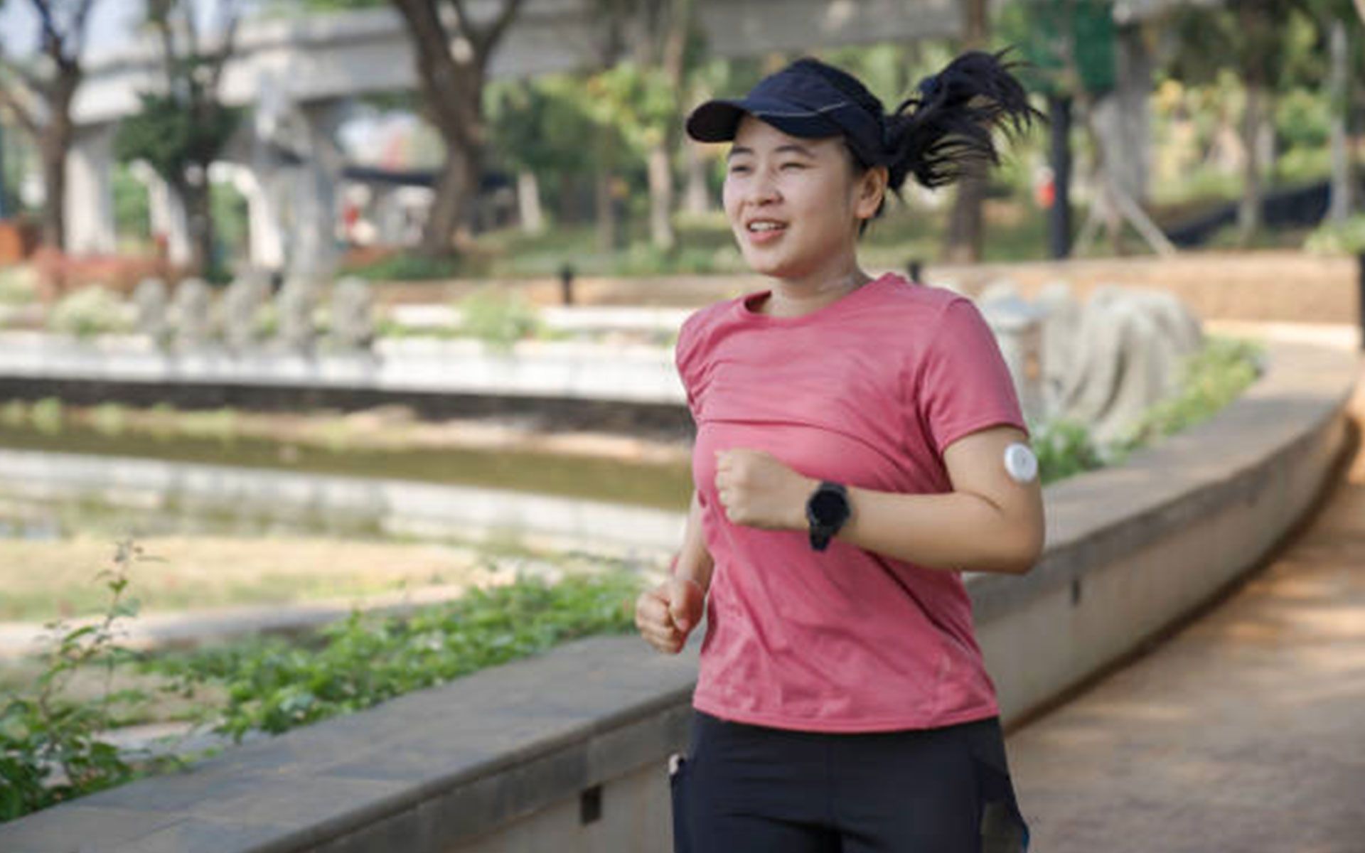 Woman jogging in a park, wearing a pink shirt and black hat, smiling, and checking her smartwatch.