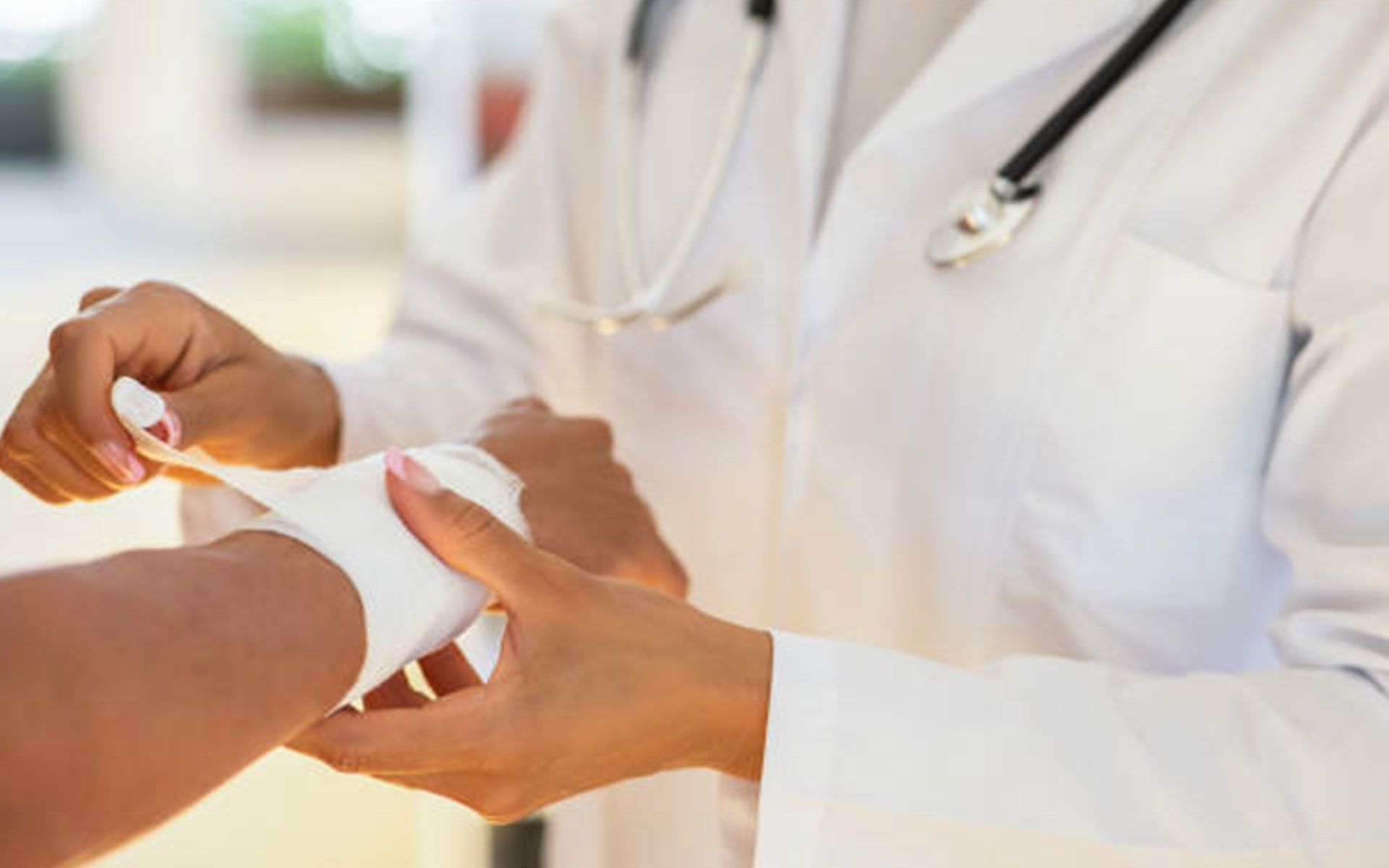 Doctor wrapping a patient's wrist with a white bandage, wearing a white lab coat and a stethoscope.