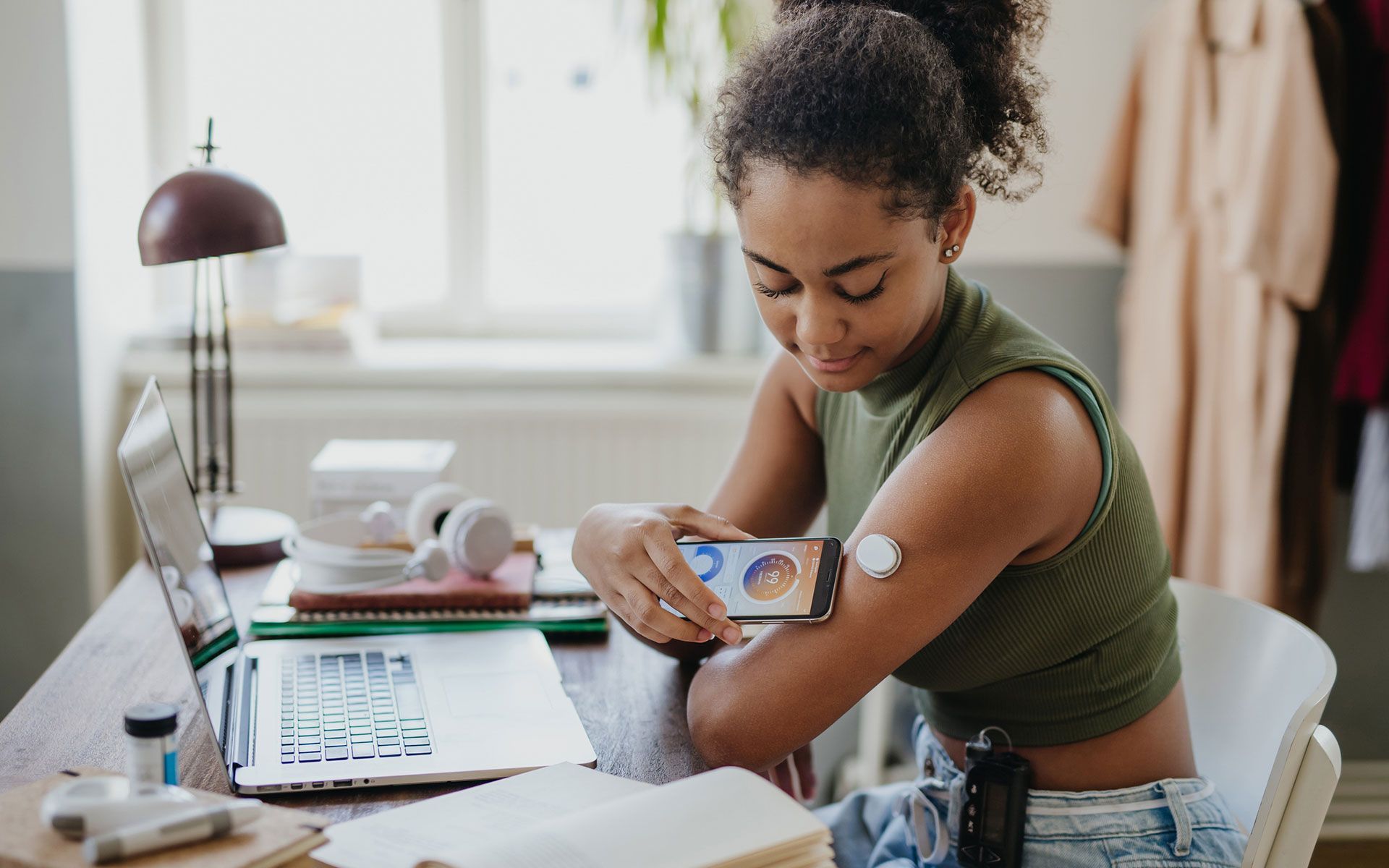Woman checks glucose levels using her phone at a desk with a laptop and medical supplies.