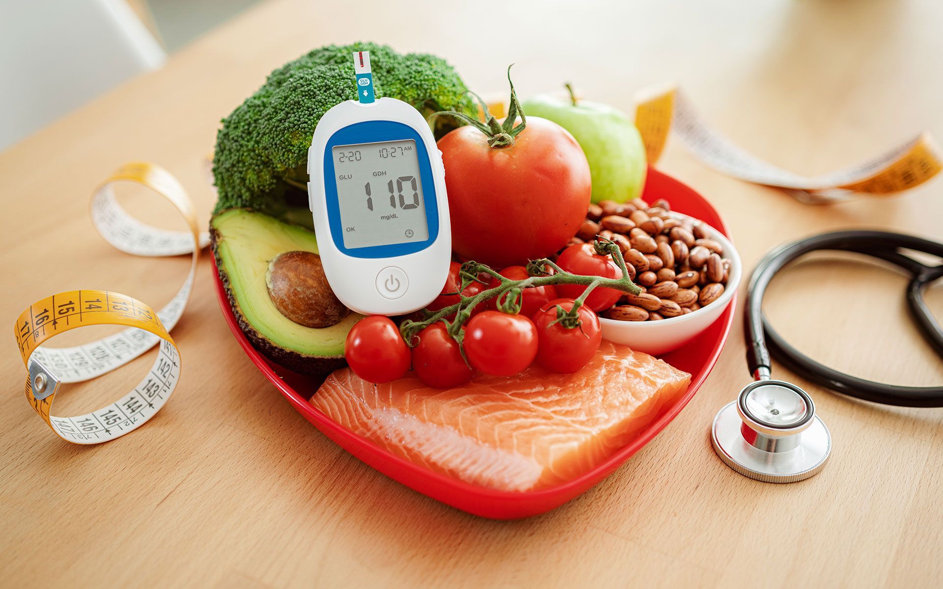 Heart-shaped plate with healthy food and glucose monitor; avocado, salmon, broccoli, tomatoes, and stethoscope.