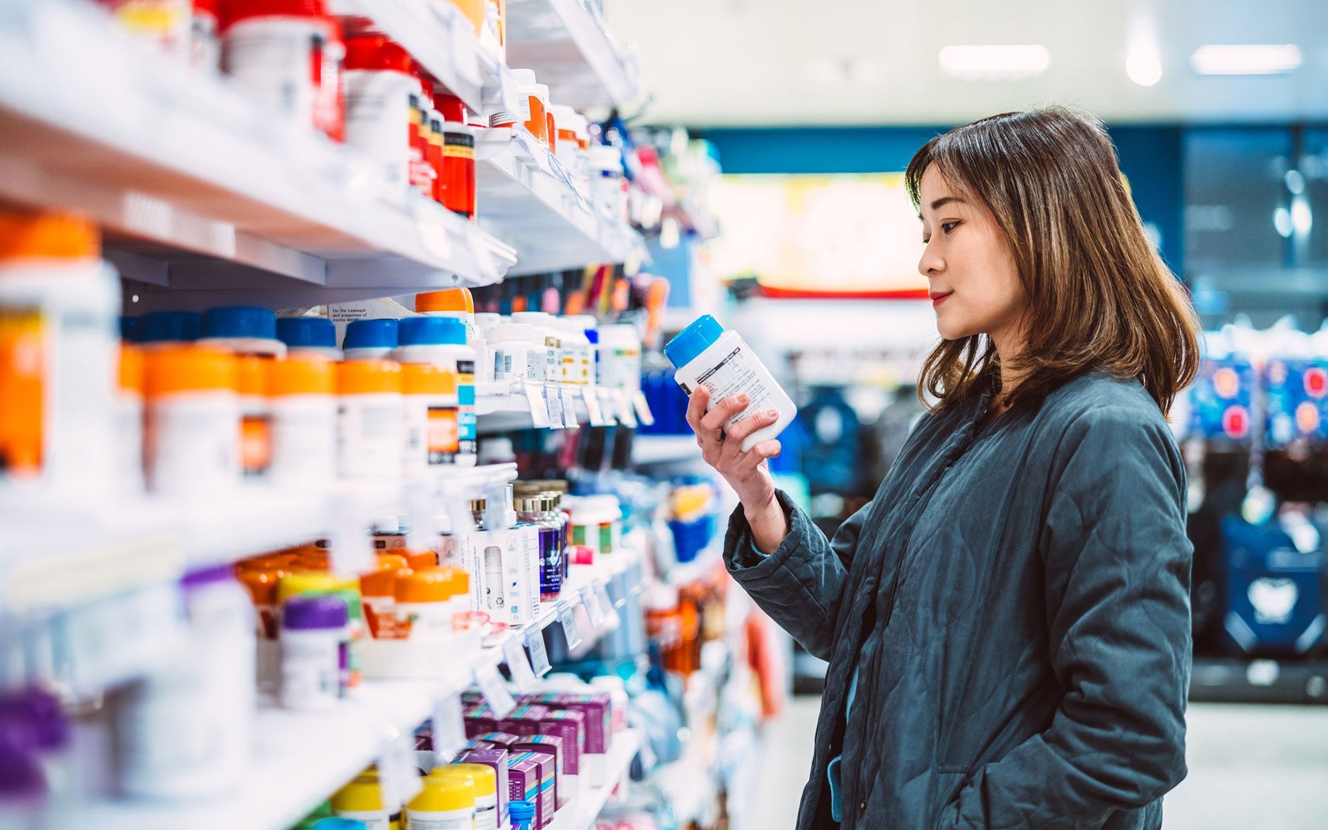 Woman in a store aisle looks at a supplement bottle, shelves filled with colorful bottles in a pharmacy.