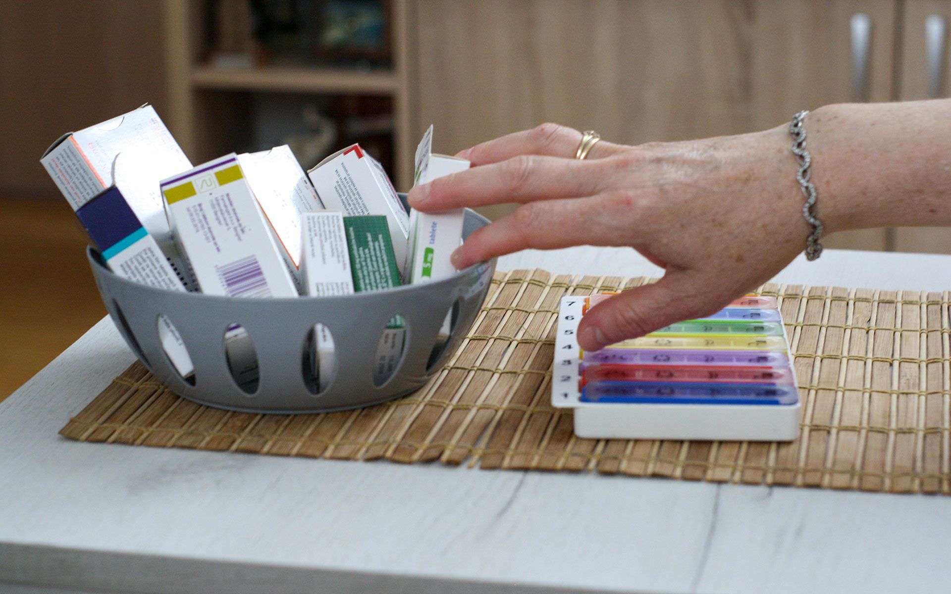 A hand reaching for a pill organizer on a table with a basket of medicine.