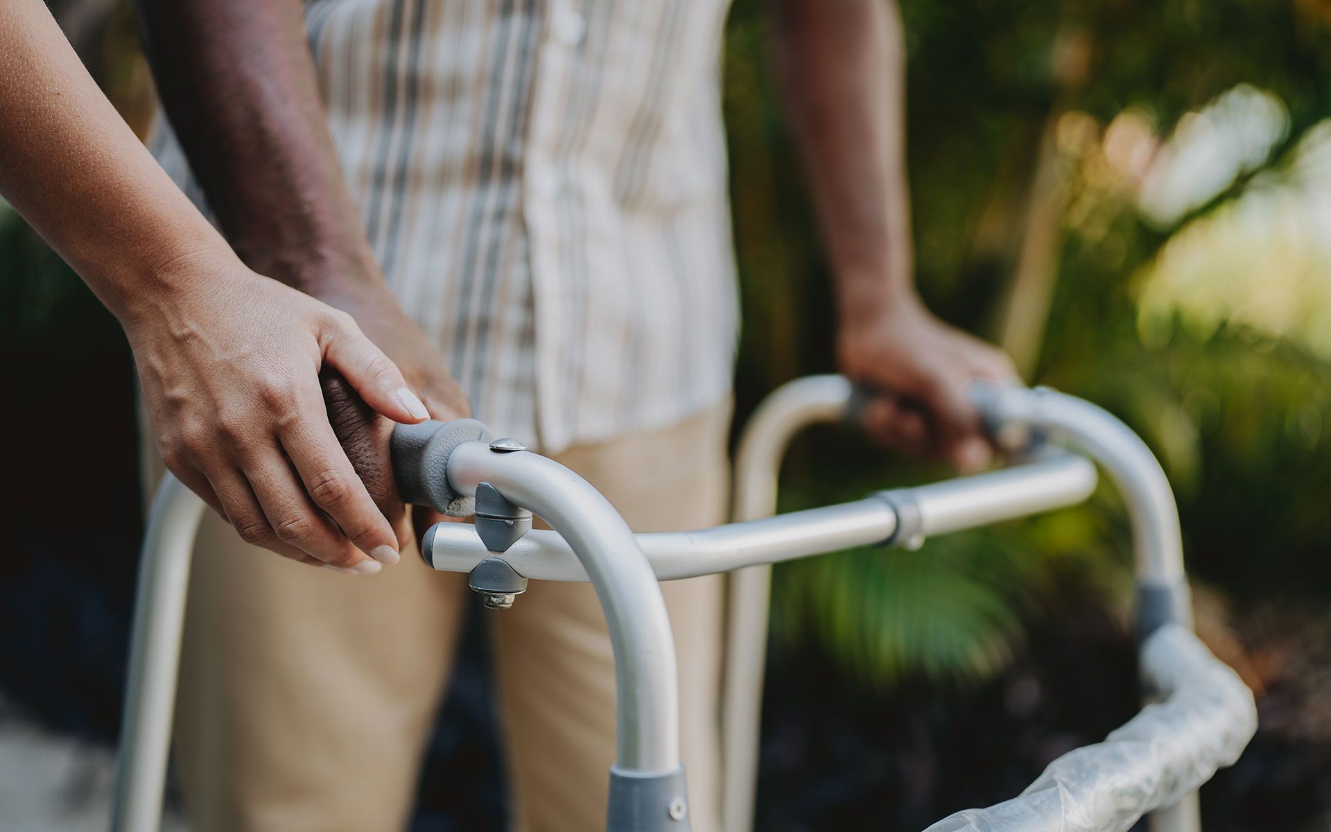 Person using a walker with another person's hands assisting. Beige clothing, outdoors.