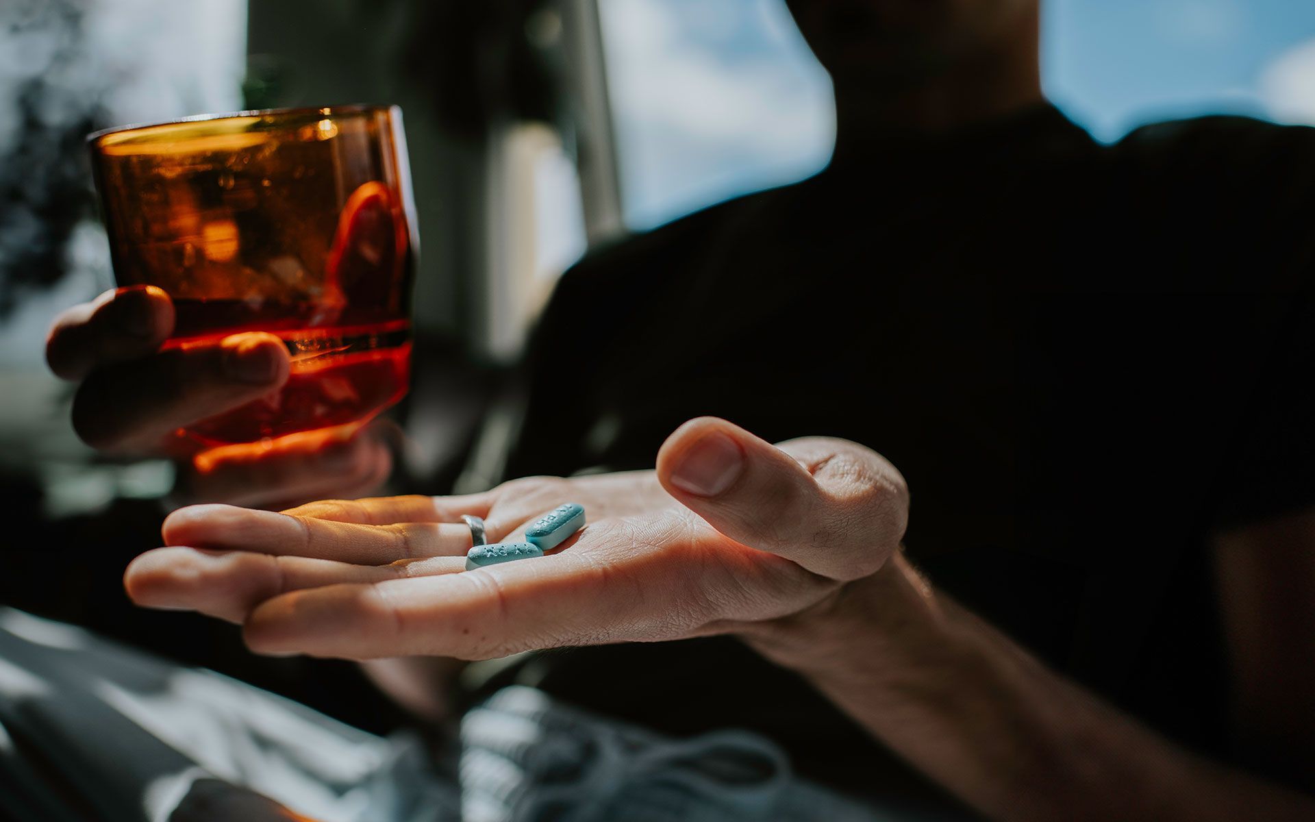 Person holding pills in one hand and a glass of amber liquid in the other.