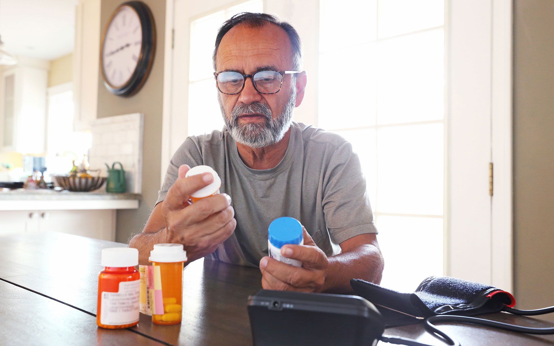 Man with glasses examining pill bottles, sitting at a table with a blood pressure monitor in a kitchen.
