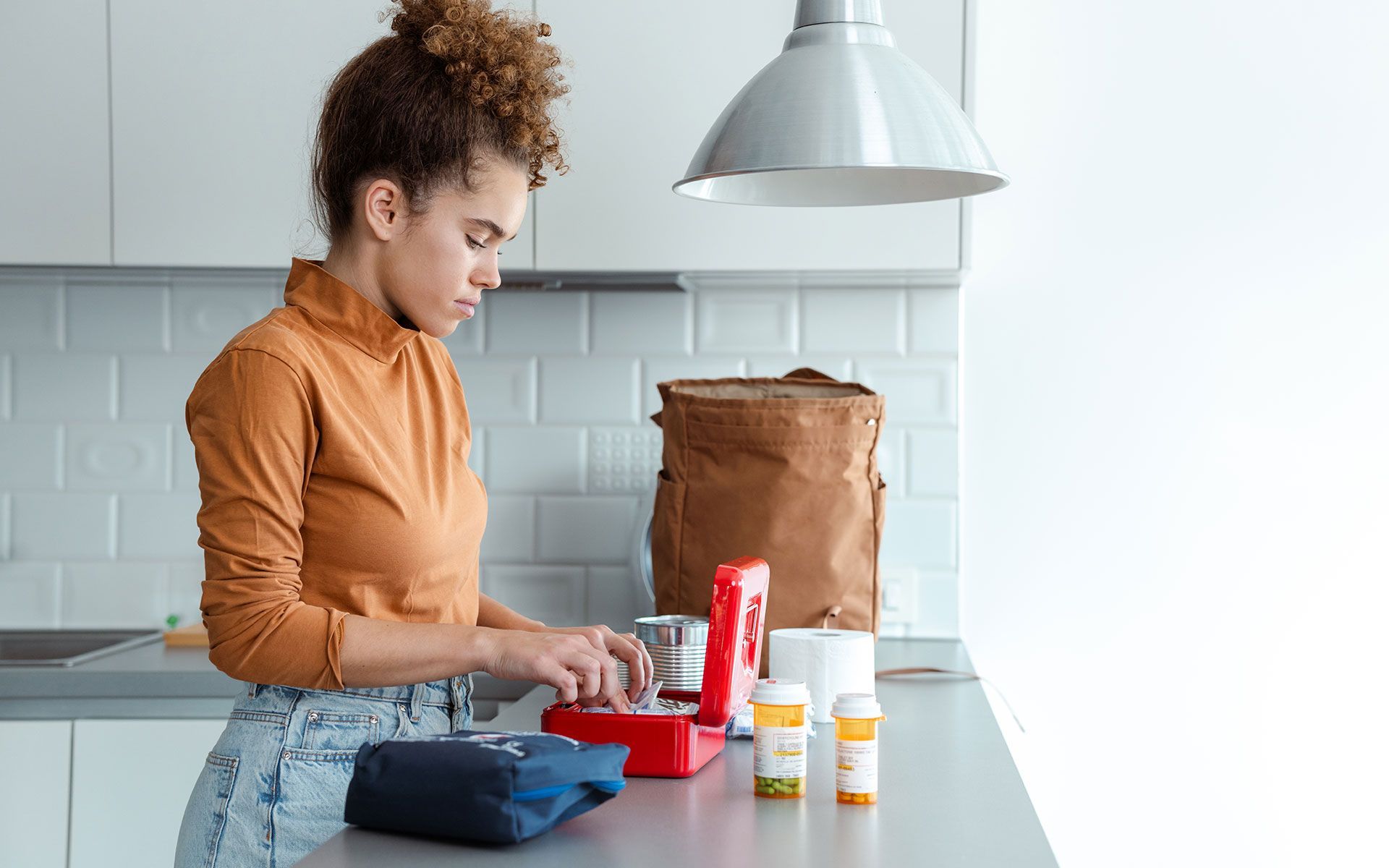 Woman preparing lunch, kitchen counter. Brown shirt, blue jeans. Pills, bag, container on counter.