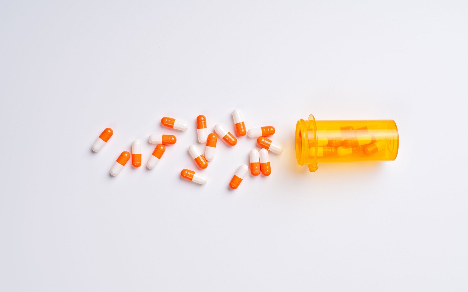 Orange and white capsules spilled from a yellow prescription bottle on a white surface.