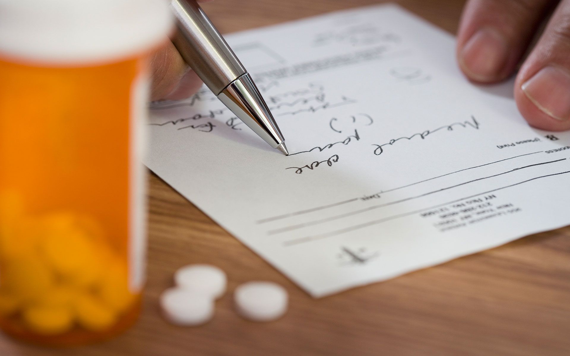 A hand writing on a prescription, with a pill bottle and pills on a wooden surface.