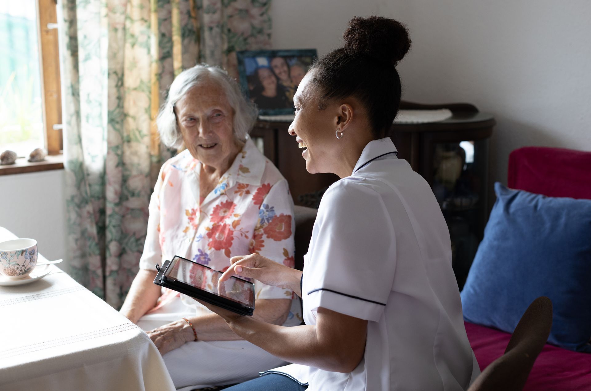 Caregiver showing a tablet to an elderly woman seated at a table. They are both smiling inside a home.