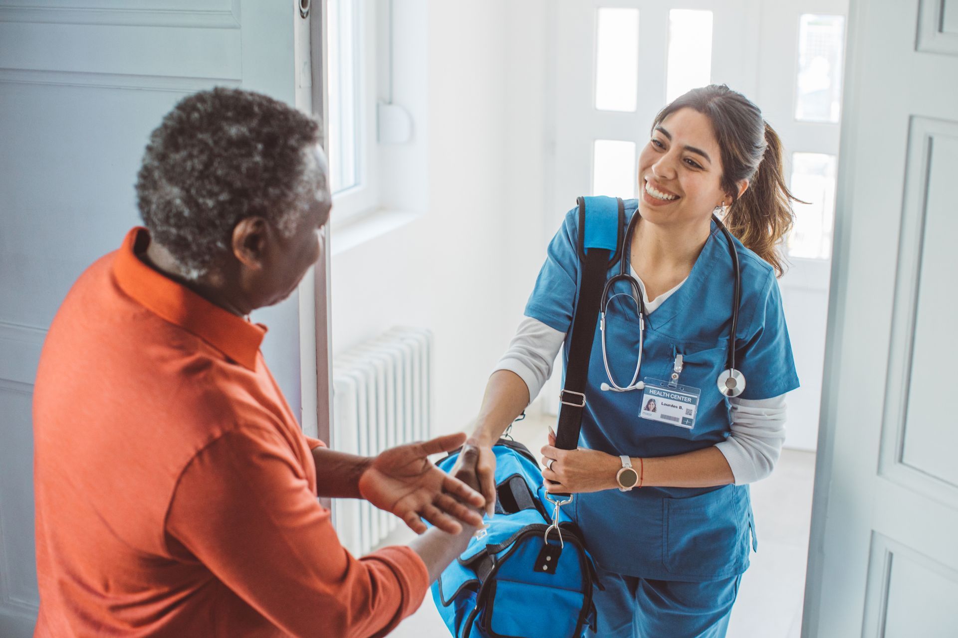 Nurse greets a smiling elderly man at his doorway. She wears scrubs and holds a medical bag.