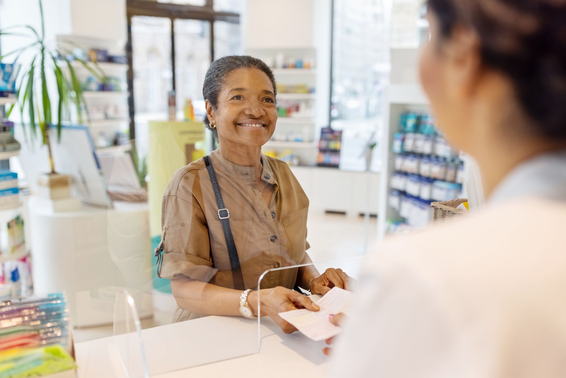 Woman smiling, handing prescription to pharmacist in brightly lit pharmacy.