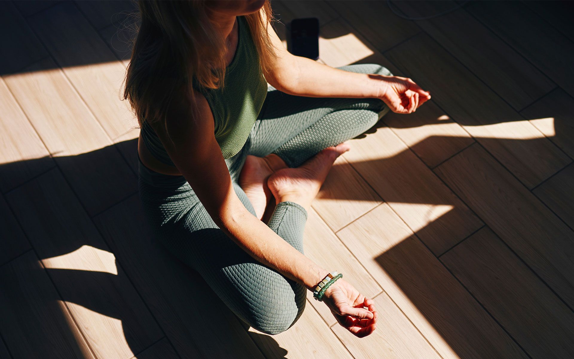 Woman sitting cross-legged on wooden floor, meditating in sunlight.