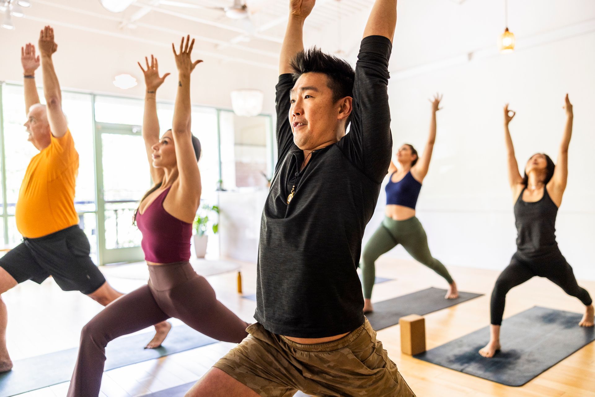 People in a yoga class doing warrior pose with arms raised. Bright studio.