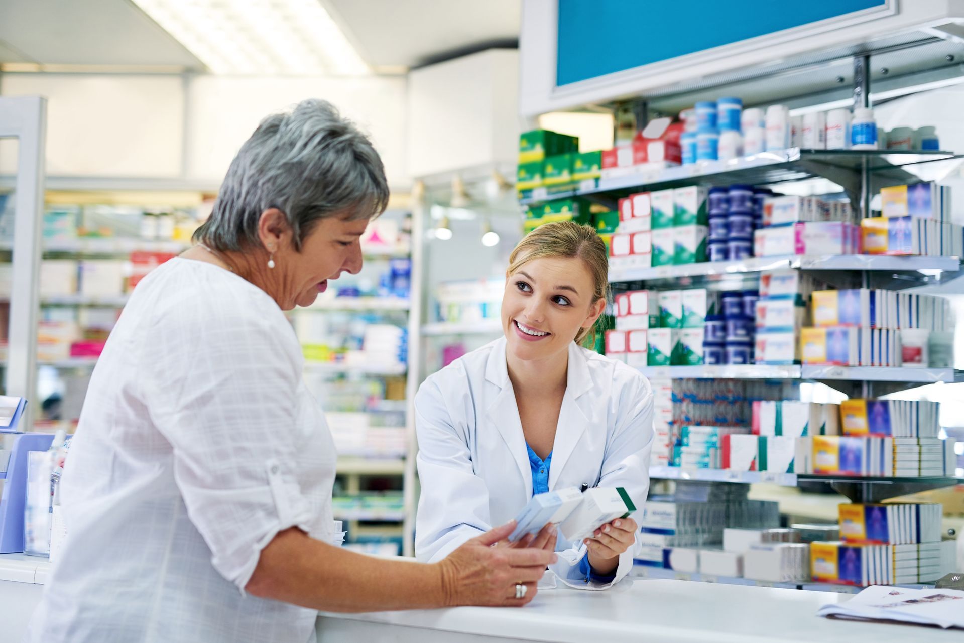 A pharmacist helps a senior woman at a pharmacy counter, smiling, holding a prescription bottle.