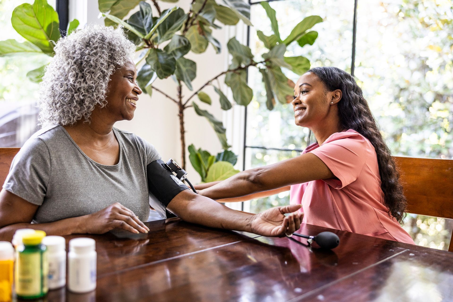 A young woman checks an older woman’s blood pressure at a table, smiling.