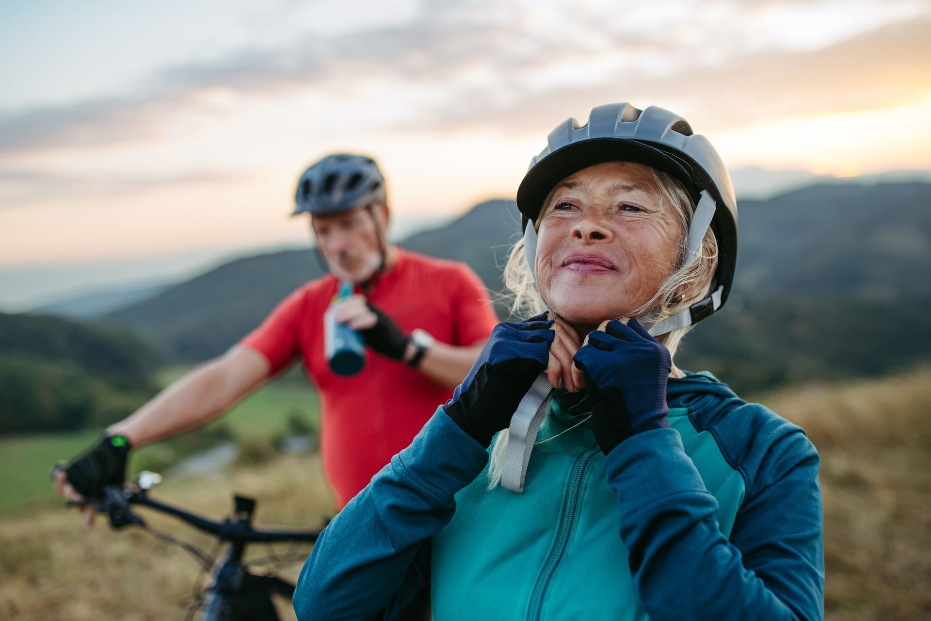 Older woman adjusting her helmet, mountain biking with a man, outdoors.