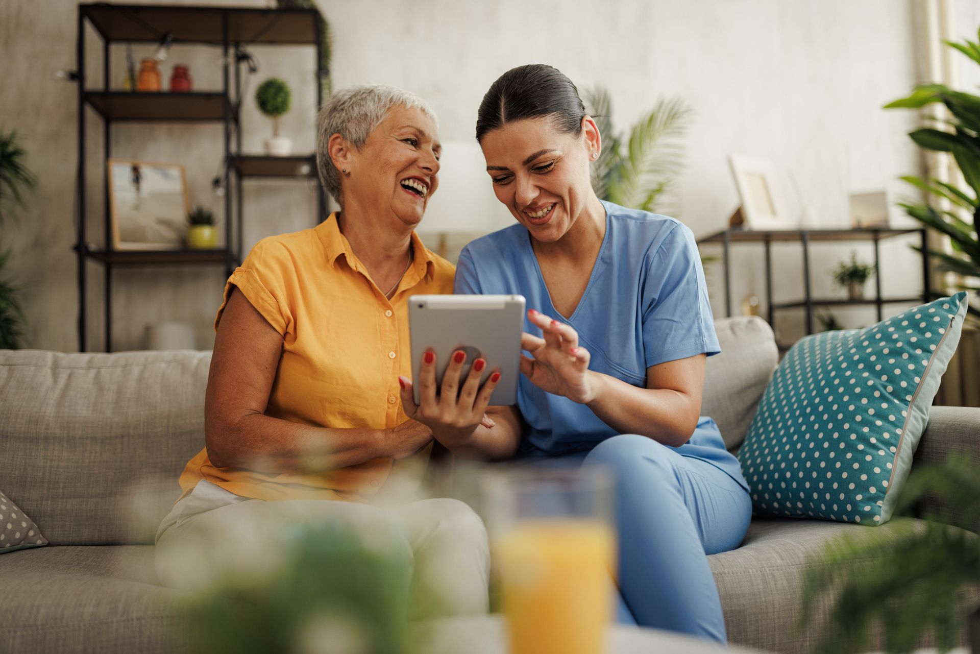 Woman and caregiver laughing together while looking at a tablet on a sofa.