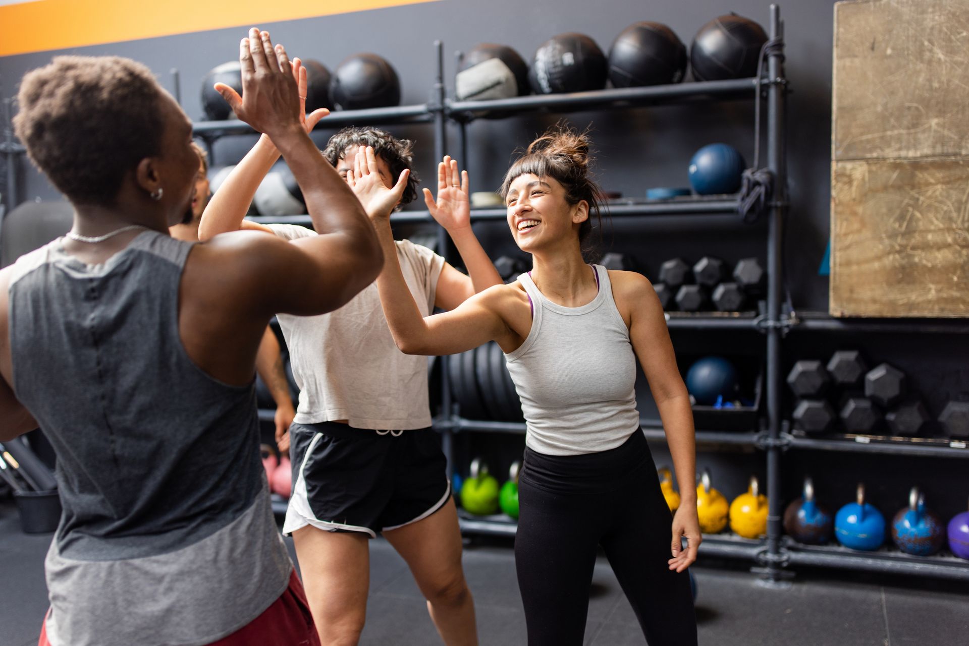 Three people in a gym high-fiving. Woman smiles, gym equipment in the background.
