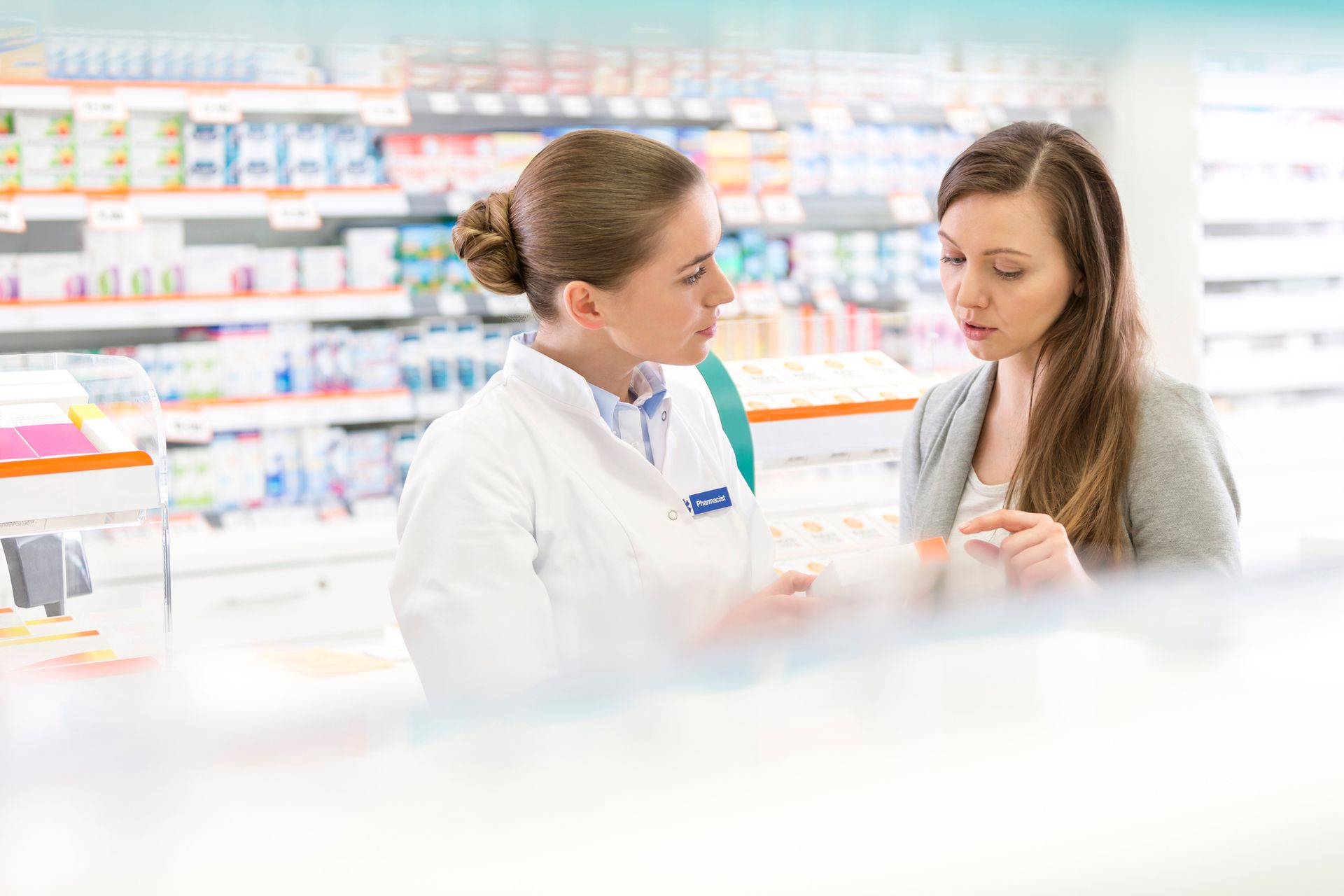 Pharmacist in white coat advises a woman in a gray jacket at a pharmacy counter.