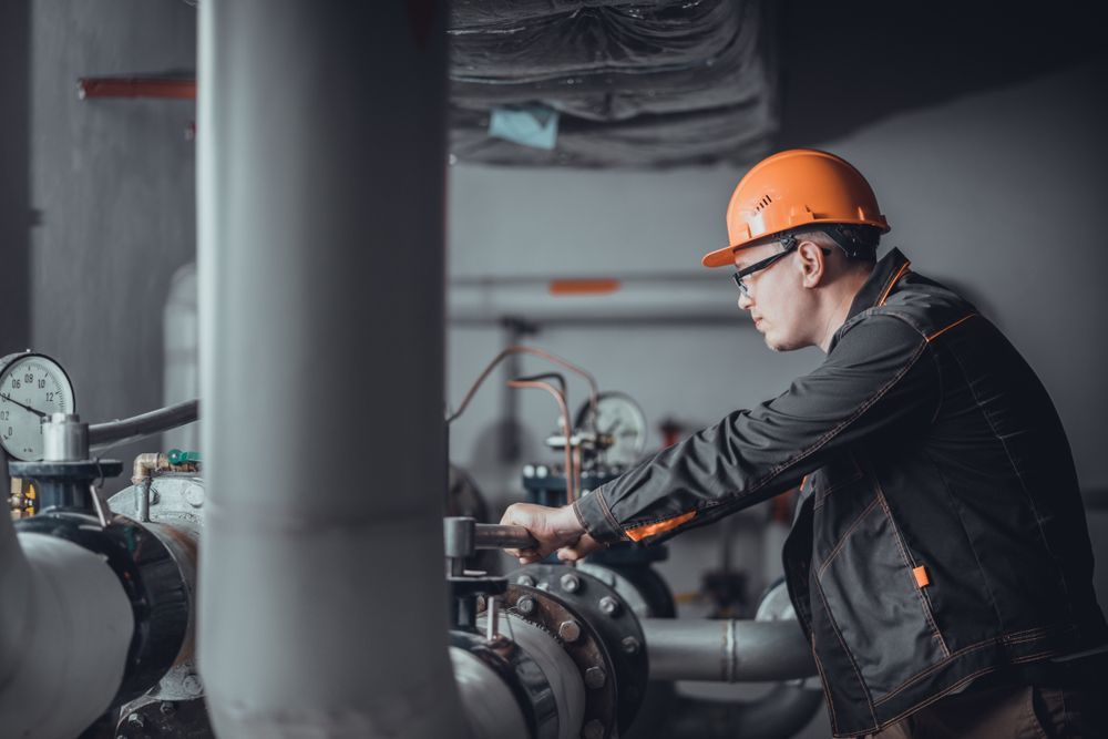 A Man In A Hard Hat Is Working On A Pipe In A Factory — Warrnambool Emergency Plumbing in Port Fairy, VIC
