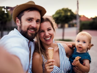 Family selfie: smiling parents with a baby eating ice cream outdoors.