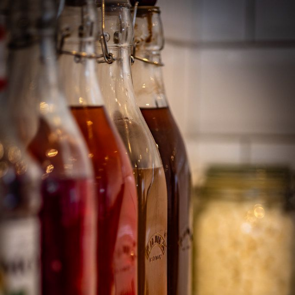 Bottles of colorful liquids in a row on a shelf, with a jar of food in the background.