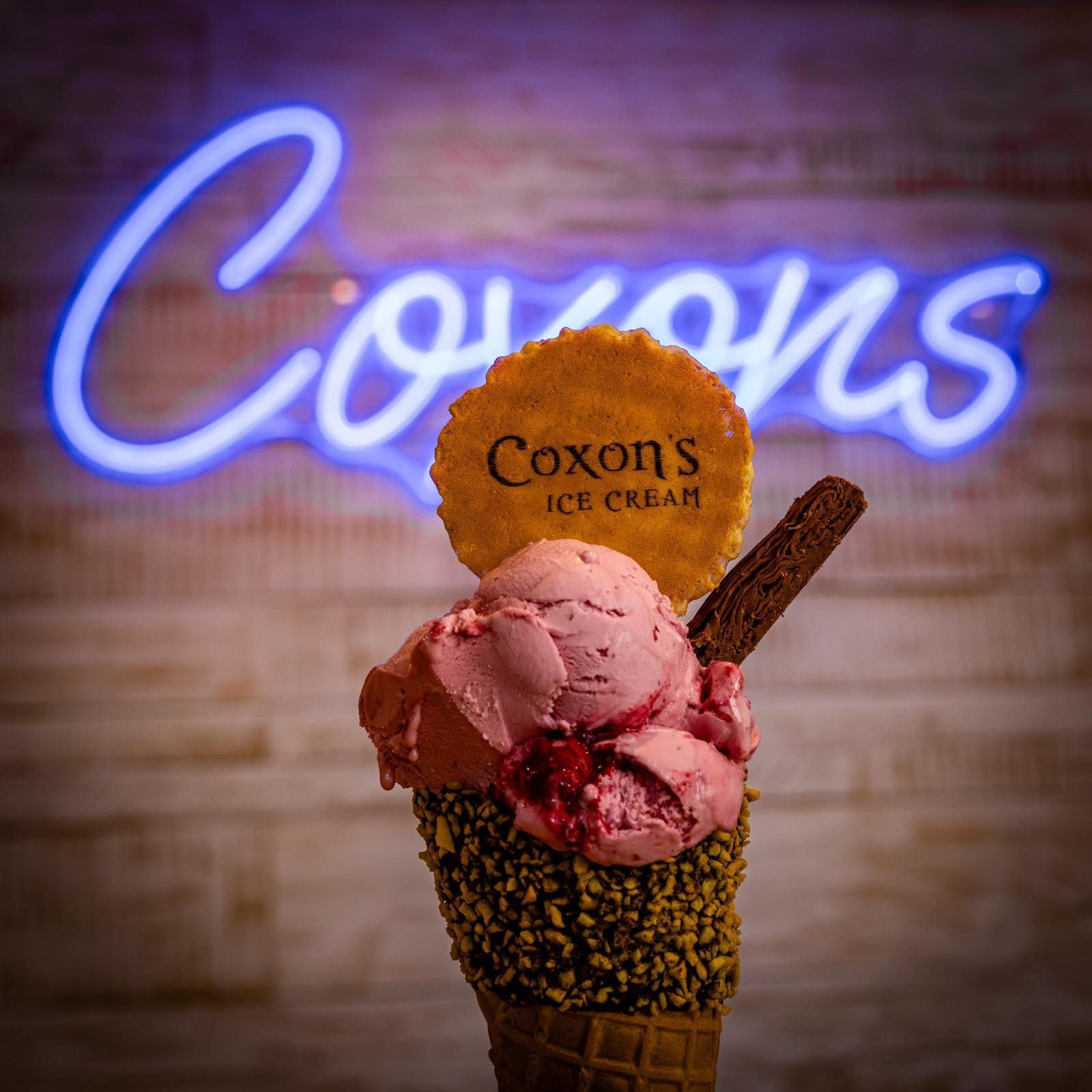 Ice cream cone with pink scoops, topped with a cookie and chocolate stick, in front of a neon 