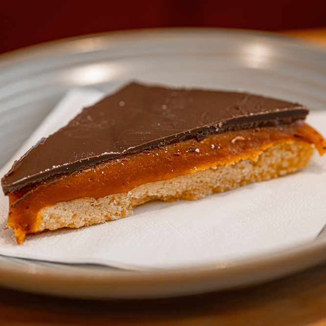 Slice of caramel shortcake with chocolate topping on a napkin, served on a grey plate.