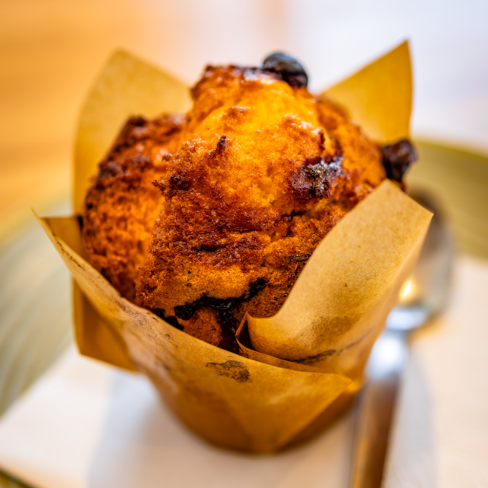 A blueberry muffin in a brown paper cup, on a plate with a spoon.