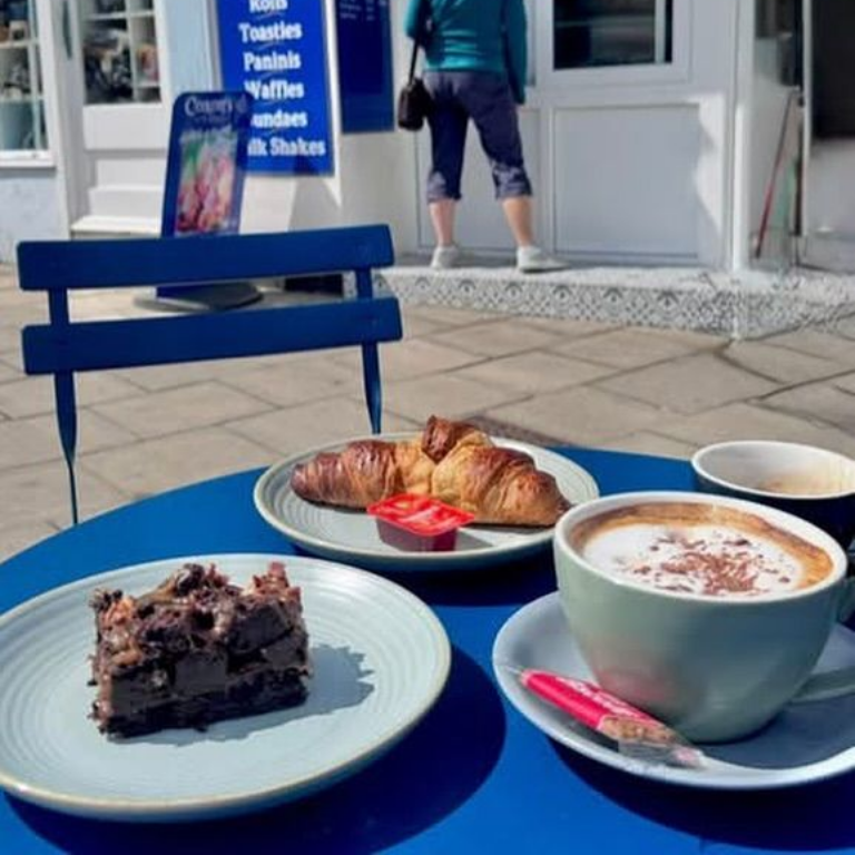 Outdoor cafe setting: coffee, pastries, and a brownie on a blue table; person in background.