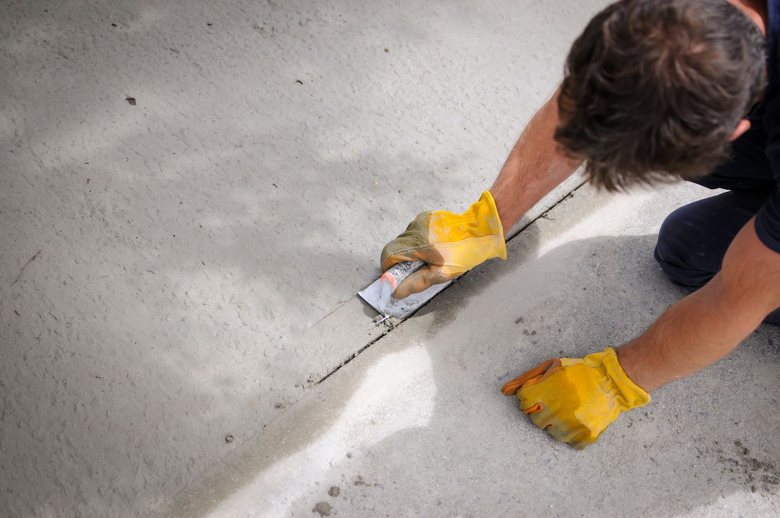 A man wearing yellow gloves is kneeling down and using a spatula on a concrete floor.
