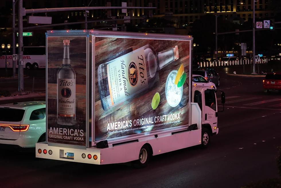 A truck with a bottle of vodka on the back is driving down a street at night.
