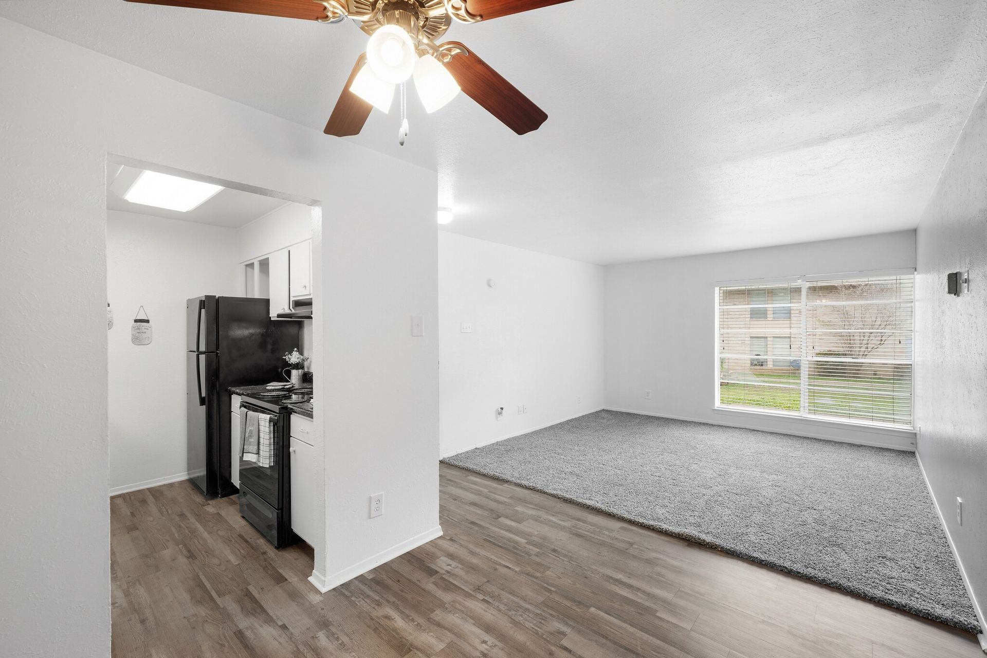A living room with a ceiling fan and a kitchen in the background.
