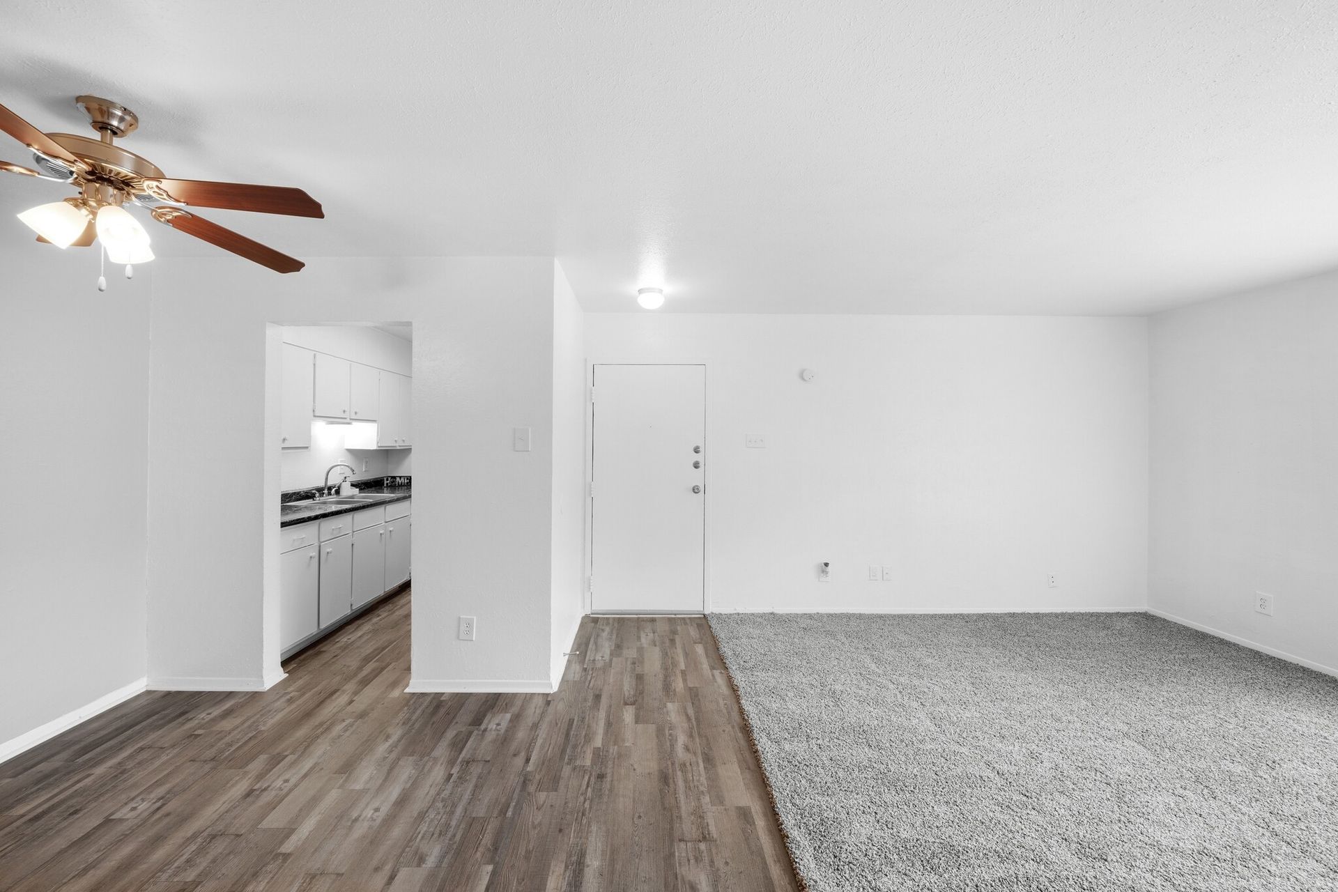 A living room with a ceiling fan and a kitchen in the background
